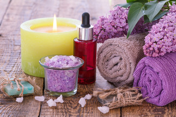 Fototapeta na wymiar Soap, burning candle, bowl with sea salt, red bottle with aromatic oil, lilac flowers and towels on wooden background