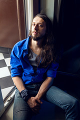 man with long hair and beard sits at home on a chair by the window