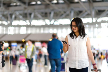 Travelers smart asian woman walking with a luggage and using smart phone  at airport terminal with blurred crowd of travelling people in background. Traveling concept.
