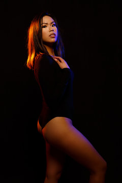 Fit young asian woman in black one piece and necklace poses against black under  orange and blue colored lights