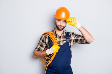 Portrait of virile harsh electrician with bristle in overall, shirt, having rolled cable on shoulder, holding hand on hardhat, standing over grey background