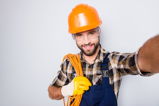 Self portrait of attractive joyful man in hardhat, overall, shirt, holding rolled cable on shoulder, blogger shooting selfie on front camera, isolated on grey background, having leisure