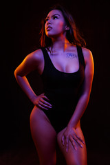 Fit young asian woman in black one piece poses against black under  orange and blue colored lights