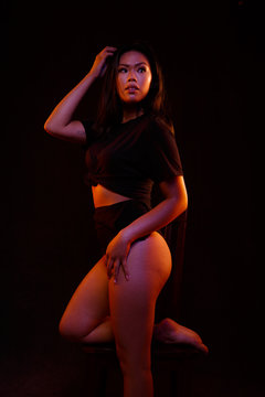 Fit young asian woman in black one piece poses on a chair under colored lights