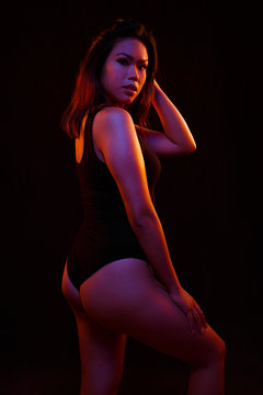 Fit young asian woman in black one piece poses against black under orange and blue colored lights
