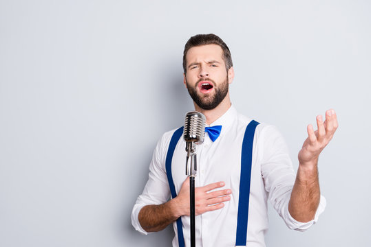 Portrait of talented attractive singer with bristle stubble in elegant classic outfit, singing hit with open mouth in microphone gesture with hand isolated on grey background