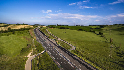 Italia, Maggio 2018 - Vista aerea dell'autostrada con automobili e camion che attraversa le colline con i campi di grano non ancora maturo