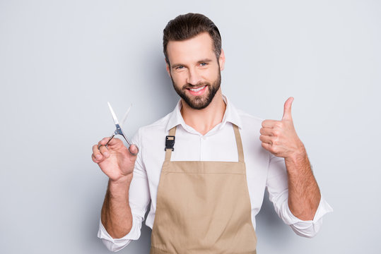 Portrait of cheerful positive barber with stubble in shirt having scissors in hand showing thumb up approve sign with finger, looking at camera, isolated on grey background