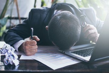 Stressed businessman in dark suit sitting at office desk full with books and papers being...