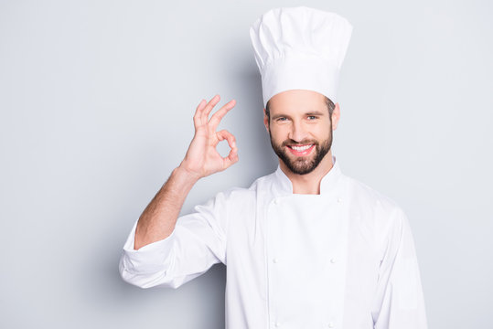 Portrait of cheerful joyful chef cook in beret and white outfit with stubble looking at camera showing ok sign, approve, advice menu isolated on grey background