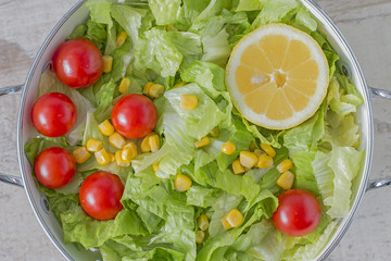 A colander with green salad, cherry tomatoes, corn and lemon. From above.