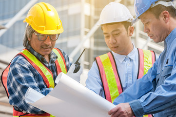 Asian civil engineer team wearing safety helmet and uniform using blueprint to meeting, discussing brain storm for solving their problem situation in middle of city town