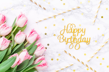 Pink tulips and gold Happy birthday letters with festive straws on white marble background. Spring and celebration concept. Copy space
