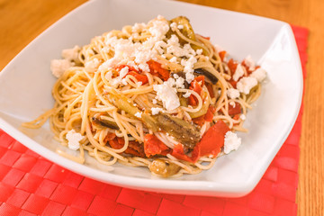 A white dish on a red place mat of with a mediterranean meal of spaghetti, feta cheese, aubergine (egg plant) and tomato