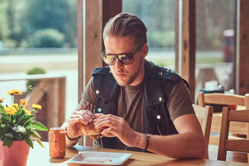 Handsome hipster with a stylish haircut and beard sits at a table, decided to dine at a roadside cafe, eating a hamburger.