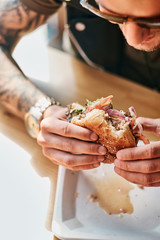 Portrait of a hungry hipster guy with a stylish haircut and beard sits at a table, decided to dine at a roadside cafe, eating a hamburger.