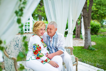 Married couple in embroidered shirts, sitting on sofa in white gazebo, on garden background