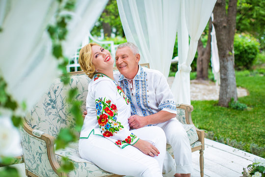 Elderly couple in shirts embroidered, sitting on sofa in white gazebo, on garden background