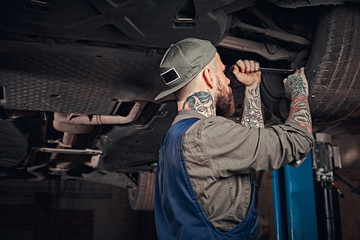 Obraz na płótnie Canvas Bearded auto mechanic in a uniform repair the car's suspension with a wrench while standing under lifting car in repair garage.