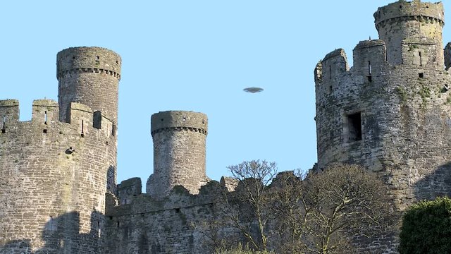 UFO sighting, Flying saucer hovering over Conwy castle, metallic reflective aircraft rotating and moving, realistic reconstruction of real UFO sighting, blue sky background.