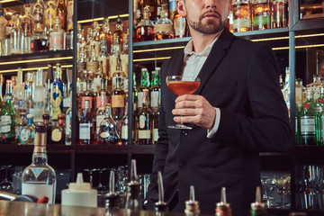 Handsome stylish male holds a glass of exclusive alcohol standing at bar counter background.