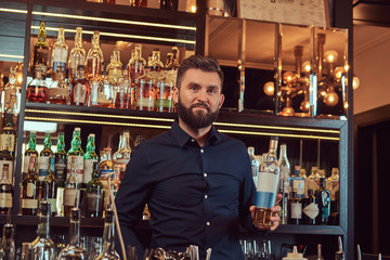 Stylish brutal bartender in a black shirt presents a bottle of exclusive alcohol at bar counter background.