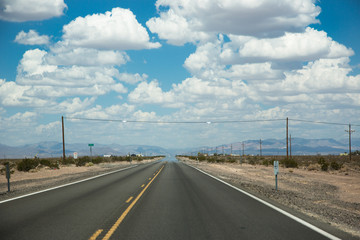 Fototapeta na wymiar Eine schnurgerade, unendliche und asphaltierte Strasse mit gelben Mittelstreifen durch die Wüste Nevadas bei blauem Himmel und dicke weissen Wolken