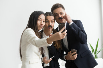Coworkers trying mustache accessory, making silly faces posing to camera while making selfie on smartphone. Colleagues having fun at work, playing childish and taking pictures during work break.