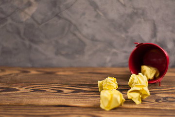 Crumpled yellow papers poured out of red metal bucket on old wooden brown table with copy space for your text. School or business concept