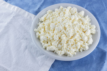 Cottage cheese on a white blue fabric background, soft cheese on a white plate, fresh homemade cheese on a white blue napkin. Dairy product in vintage style, healthy lifestyle. Copy space