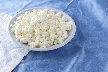Cottage cheese on a white blue fabric background, soft cheese on a white plate, fresh homemade cheese on a white blue napkin. Dairy product in vintage style, healthy lifestyle. Copy space
