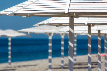 Fototapeta na wymiar White wooden umbrellas on pebble beach. Empty sea shore at sunny day