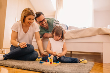Young parents and their daughter are playing with a wooden game on floor in bedroom