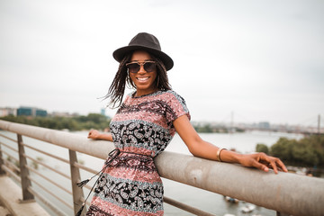 Smiling african american woman in a black hat is standing near bridge fence while getting ready for photo shooting