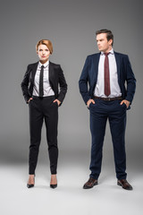 confident businesswoman and businessman standing with hands in pockets, isolated on grey
