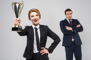 excited businesswoman with award and upset businessman behind, isolated on grey