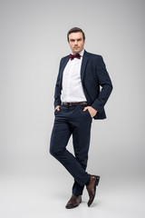 handsome man posing in tuxedo with hands in pockets, isolated on grey