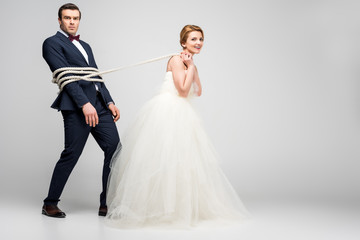 beautiful bride pulling groom bound with rope, isolated on grey, feminism concept