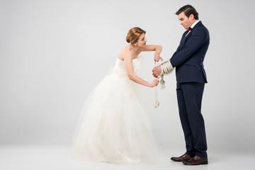 bride in wedding dress bounding groom with rope, isolated on grey, feminism concept