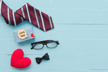 Happy fathers day concept. Red tie, glasses, gift box with LOVE DAD text and handmade red heart on bright blue pastel wooden table background.