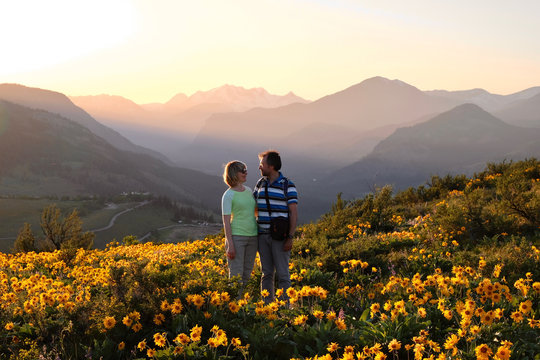 Happy friends surrounded by blooms. Smiling man and woman looking at each other on arnica meadow in bloom. Travel Pacific Northwest. Washington State. United States of America