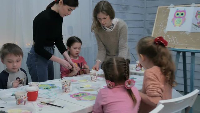 Children boys and girls sitting together around the table in classroom and drawing. With them is their young and beautiful teacher.