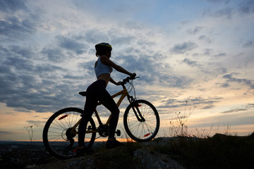 Back view athletic female cyclist in helmet with bicycle on rock under beautiful evening sky with clouds on background of city in the distance. Copy space