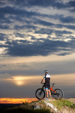 Rear view of an athletic male with a bicycle enjoying the evening sky and the beautiful sun at sunset on top of a mountain with a breathtaking landscape of hills in the distance.