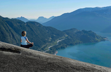 Fototapeta na wymiar Outdoor yoga retreat. Woman in lotus pose meditating on mountain top with beautiful views. Whistler. Stawamus Chief Peak. British Columbia. Canada.