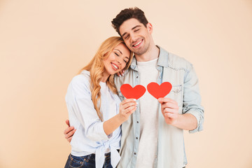 Image of smiling man and woman in love wearing denim clothing hugging together with closed eyes and holding two red paper hearts, isolated over beige background