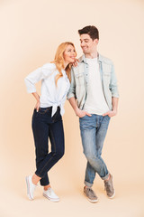 Full length image of positive caucasian couple man and woman in denim clothing smiling and looking at each other, isolated over beige background