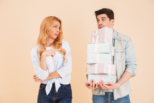 Portrait of disappointed woman pointing finger at upset man standing with lots of gift boxes, isolated over beige background