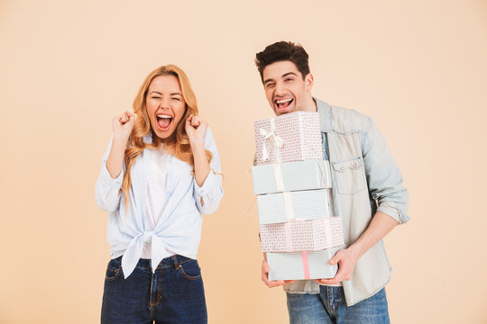 Image of surprised woman shouting in happiness while handsome man holding lots of gift boxes, isolated over beige background