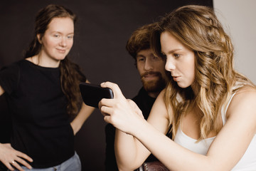 Three people in casual outfits holding smartphone and looking at photos on it while standing in studio. 